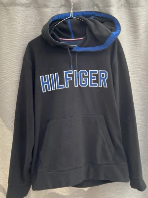 Svart Hilfiger hoodie med blå detaljer - Cool svart hoodie från Hilfiger med blå kant på huvan och stor vit/blå logotyp på bröstet. Klassisk passform, stor känguruficka och justerbar huva med snören. Perfekt för dig som gillar streetwear och vill ha något bekvämt och snyggt.