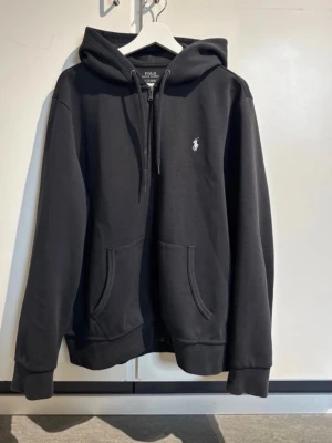 Svart hoodie från Polo Ralph Lauren - Snygg svart hoodie från Polo Ralph Lauren med dragkedja, huva och fickor framtill. Nypris: 2.195kr. Tveka inte att höra av dig vid frågor!