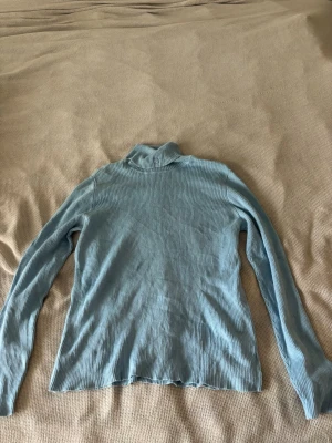 Ljusblå ribbstickad polotröja RL - Ljusblå ribbstickad polotröja från Ralph Lauren med diskret RL-logga på kragen. Tröjan har lång ärm och är tillverkad i mjukt stickat material som ger en skön passform. Perfekt för lager-på-lager och snygg att matcha med jeans.