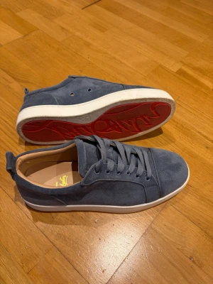 Christian Louboutin skor - Snygga sneakers i ljusblå/grå mocka från Christian Louboutin med klassisk röd sula och vita kanter. Skorna har rund tå, matchande blå skosnören och diskret logga på innersulan. Perfekta för dig som vill ha en stilren och exklusiv look. Skorna är använda endast 2 gånger och är i perfekt skick. Tyvärr finns det ingen box. Priset är ej hugget i sten och kan såklart diskuteras😁
