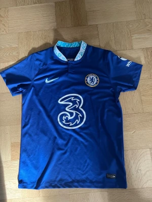 Chelsea matchtröja Kanté #7 Nike - Säljer en blå Chelsea FC matchtröja från Nike med Kanté och nummer 7 på ryggen. Tröjan har klubbmärke, Nike-logga och sponsortryck framtill samt vitblå krage med mönster. Perfekt för fotbollsfans och samlare.