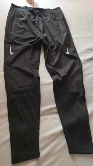 Svarta träningsbyxor från Nike - Säljer ett par svarta träningsbyxor från Nike med vita loggor på båda benen. Byxorna har dragkedjor vid fickorna, resår i midjan och ventilerande meshpaneler på benen. Perfekta för löpning eller gym. Materialet är lätt och andas.