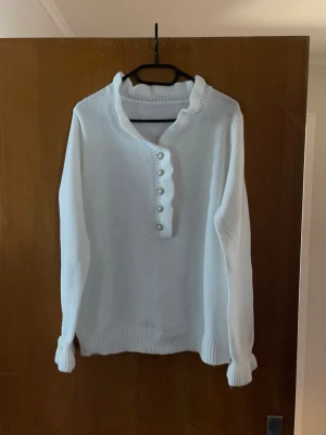Vit stickad tröja med pärlknappar - Snygg vit stickad tröja med lång ärm och ribbade muddar. Tröjan har en unik krage och dekorativa pärlknappar framtill. Perfekt för dig som gillar en stilren och söt look.