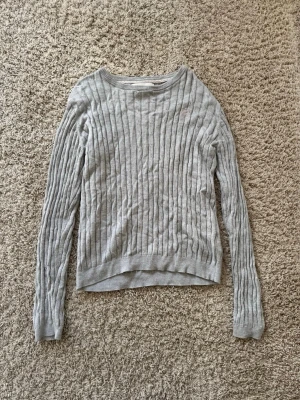 Ljusgrå ribbstickad tröja - En stilren ljusgrå stickad tröja med ribbade vertikala ränder. Tröjan har rund halsringning, långa ärmar och ribbade muddar vid ärmslut och nederkant. Perfekt för lager på lager och enkel att matcha med olika outfits.