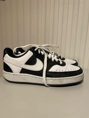 Nike svartvita låga sneakers - Snygga svartvita Nike sneakers i lågt utförande med klassisk swoosh på sidan. Skorna har vita snören, svart och vit ovandel i läder och tydlig Nike-logga på hälen. Perfekta för dig som gillar stilrena och tidlösa sneakers.