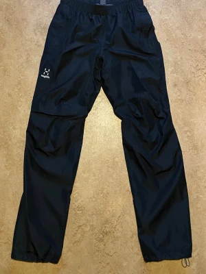 Haglöfs Mila Rain Pant Men - Som nya! - Snygga svarta täckbyxor från Haglöfs med resår i midjan och justerbara benslut. Tillverkade i slitstarkt, vattenavvisande material som passar perfekt för kalla och blöta dagar. Diskret logga på vänster lår och enkel, rak passform.