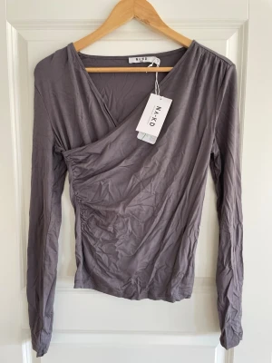 Grå omlottopp från NA-KD S - Snygg grå omlottopp från NA-KD med v-ringning och långärmad design. Toppen har en cool omlott-effekt och lätt rynkade detaljer på sidan. Perfekt för dig som gillar stilrena och trendiga plagg. Materialet känns mjukt och lätt.