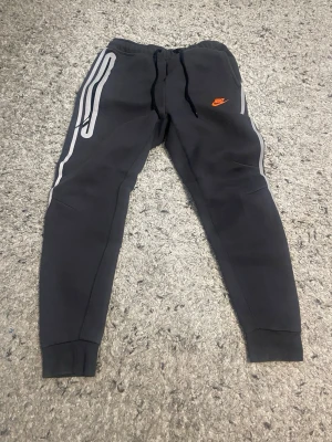 Svarta Nike mjukisbyxor med tryck - Säljer ett par svarta mjukisbyxor från Nike med vita detaljer längs benen och orange Nike-logga på vänster sida. Byxorna har dragsko i midjan och muddar vid bensluten. Perfekta för chill eller träning.