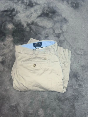  Polo Ralph Lauren Chinos Shorts – Beige - stilrena chinos-shorts från Polo Ralph Lauren i en perfekt beige nyans. Modellen är "Stretch Classic Fit", vilket gör dem extremt bekväma samtidigt som de håller formen. Perfekta för sommarens alla tillfällen! • Märke: Polo Ralph Lauren • Passform: Stretch Classic Fit (Skön passform med lite stretch). Storlek w36 L/XL • Detaljer: Broderad logga ovanför bakfickan, snygga knappar och hög kvalitet i tyget. • Skick: Toppskick, nytvättade och redo för soliga dagar. Ett måste i sommargarderoben 