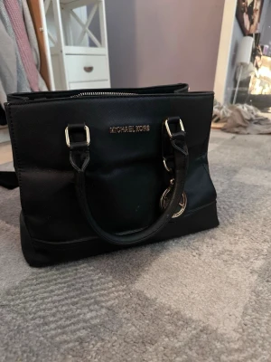 Svart handväska från Michael Kors - Stilren svart handväska från Michael Kors (är inte helt säker på att den är äkta) med guldiga detaljer och logotyp. Väskan har två rejäla handtag och en klassisk, fyrkantig form. Materialet är strukturerat och slitstarkt, perfekt för dig som gillar trendiga accessoarer.