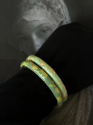 Grönt armband med gulddetaljer - Säljer ett stilrent armband i ljusgrön färg med eleganta guldfärgade blommönster. Armbandet är hårt och har en rund form, tillverkat i metall med emaljfinish. Perfekt för dig som vill ha något unikt och färgglatt till din outfit.