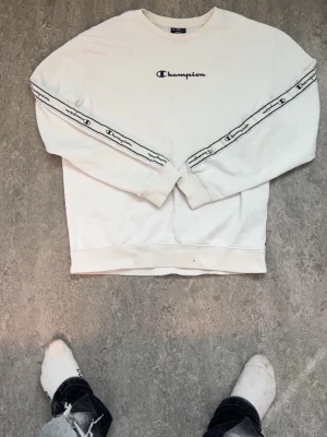Vit sweatshirt från Champion - Vit sweatshirt från Champion med svart logotyp på bröstet och logoband längs ärmarna. Rund halsringning och ribbade muddar. Perfekt för en avslappnad och sportig look.