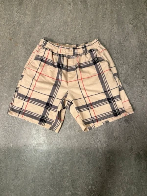 Beige rutiga shorts med resår - Säljer ett par beige shorts med klassiskt rutigt mönster i svart och rött. De har elastisk midja för skön passform och är perfekta för varma dagar. Stilen är avslappnad och trendig, passar dig som gillar att sticka ut lite extra.