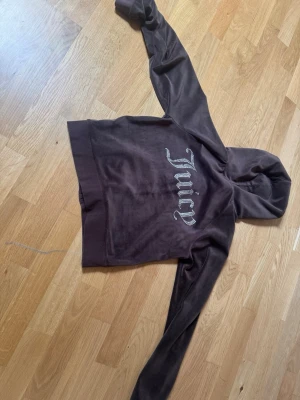 Brun Juicy Couture velour hoodie - Mjuk brun hoodie i velour från Juicy Couture med dragkedja, fickor fram och huva med snören. På ryggen står det 'Juicy' i glittriga stenar och på bröstet finns en liten logga. Perfekt för en avslappnad och trendig look.