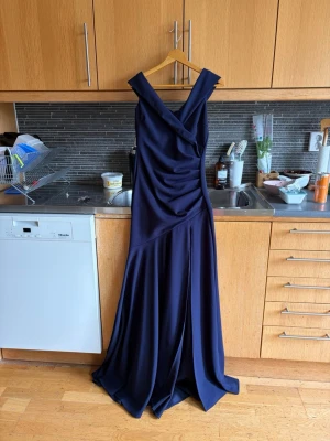 Marinblå långklänning med v-ringning - Elegant marinblå långklänning med v-ringad omlott-design och breda axelband. Klänningen har draperad effekt framtill och ett flödande, svepande tyg som ger en lyxig känsla. Perfekt för speciella tillfällen och bal. Nyskick med lapp kvar.XS men passar upp t M