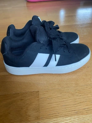 Svarta sneakers  - Snygga svarta sneakers med klassiska vita ränder på sidan och vit sula. Skorna har låga skaft, tjocka svarta skosnören och en mjuk vadderad insida. De är helt nya och oanvända 