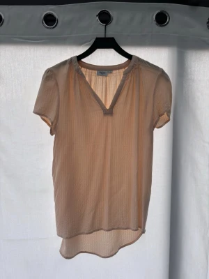 Beige blus från Saint Tropez - Säljer en luftig beige blus från Saint Tropez med korta ärmar och v-ringning. Blusen har ett subtilt vågigt mönster i tyget och är något längre baktill. Perfekt för en avslappnad och stilren look.