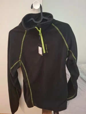 Svart fleece från Stadium med dragkedja - Svart fleece-tröja från Stadium med limegröna sömmar och halv dragkedja framtill. Tröjan har hög krage och är tillverkad i mjukt fleecematerial, perfekt för lager-på-lager. Snygg detalj med kontrastsömmar och dragkedja i neonfärg.