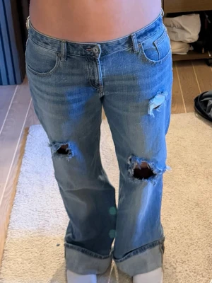 Blå slitna wide jeans från Zara  - Säljer ett par blå bootcut jeans med flera stora slitningar och hål på benen för en riktigt trendig och avslappnad look. Jeansen har klassisk femficksdesign och normal midja. 