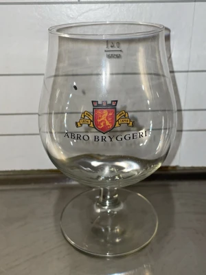 Glas från Åbro Bryggeri - Säljer ett glas från Åbro Bryggeri med bryggeriets logga och text på framsidan. Glaset är genomskinligt med rundad kupa och stabil fot. Perfekt för dryckesentusiasten eller samlaren av bryggeriprylar.