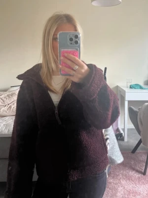 Mörkröd teddyjacka med dragkedja - Mysig teddyjacka i mörröd färg med dragkedja framtill och hög krage. Jackan har en avslappnad passform och är tillverkad i fluffigt teddy-material som håller dig varm. Perfekt för kyliga dagar och enkel att styla till jeans eller leggings.