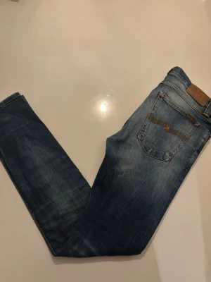 Blå jeans från Nudie Jeans - Snygga blå jeans från Nudie Jeans med klassisk femficksdesign och gul kontrastsöm. Jeansen har en lätt tvättad look och Nudie-loggan broderad på bakfickan. Perfekta för en avslappnad och trendig stil. Storlek 32-32!
