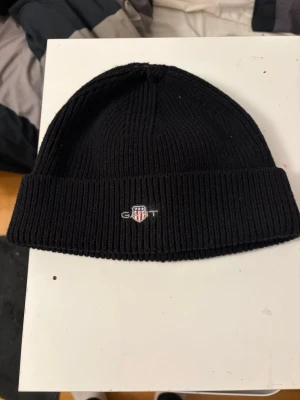 Svart ribbstickad mössa från Gant - Säljer en svart ribbstickad mössa från Gant med uppvikt kant och liten broderad logga framtill. Klassisk och enkel design som passar till det mesta. Tillverkad i mjukt material som håller värmen under kalla dagar.