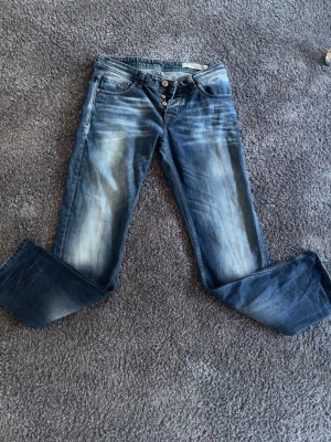Blå bootcut Jeans 31/32 - Snygga blå bootcut jeans från Replay med slitningar och ljusare partier framtill och baktill. Klassisk femficksmodell med knappgylf och läderpatch bak i midjan. Perfekta för en avslappnad och trendig look.
