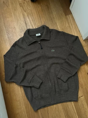Brun ziptröja från Lacoste - Mörkbrun ziptröja från Lacoste med klassisk krage och ribbade muddar. Tröjan har dragkedja framtill och den ikoniska gröna krokodilloggan på bröstet. Perfekt till lager på lager och enkel att matcha med jeans.🐊