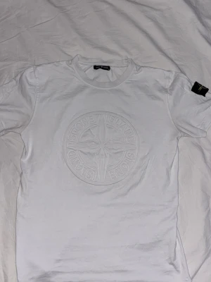 Vit Stone Island t-shirt med logga - Vit t-shirt från Stone Island med stor, ton-i-ton broderad logga på bröstet och klassisk patch på ärmen. T-shirten har rund hals och korta ärmar. Perfekt för dig som gillar stilrena och exklusiva tröjor. DU FÅR MED EN POLO MÖSSA PÅ KÖPET( gratis)