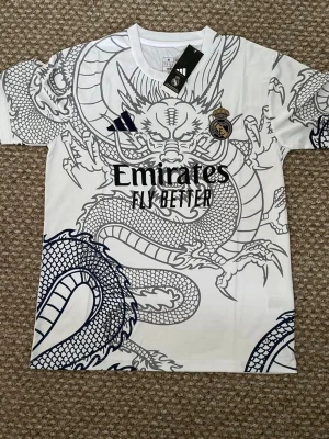 Real Madrid x Adidas draktröja - Snygg vit Real Madrid-tröja från Adidas med en stor svart drakdesign över hela framsidan. Tröjan har klubbmärke, Adidas-logga och Emirates Fly Better-tryck på bröstet. Perfekt för dig som vill sticka ut på planen eller på läktaren.