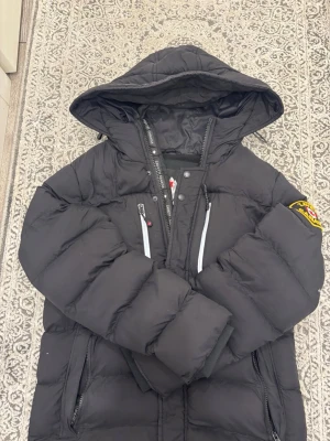 Svart jumper från Zavetti Canada - Snygg svart jumper från Zavetti Canada med huva och flera dragkedjeförsedda fickor. Jackan har en patch på ärmen och coola vita detaljer vid bröstfickorna. Perfekt för kalla dagar och riktigt skön att bära.