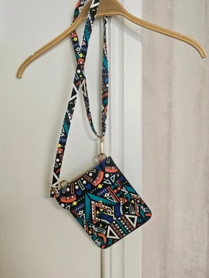 Färgglad axelväska med mönster - Säljer en liten axelväska med färgstarkt geometriskt mönster i blått, orange, vitt och svart. Väskan har justerbar axelrem och magnetlås. Materialet är konstläder och insidan har ett huvudfack. Perfekt för dig som gillar unika accessoarer!