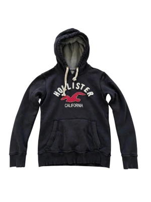 Svart hoodie från Hollister  - Svart hoodie från Hollister med stor vit logga och röd fågel på bröstet. Mjuk insida, huva med beige dragsko och magficka framtill. Perfekt för en avslappnad och cool stil. I storlek S