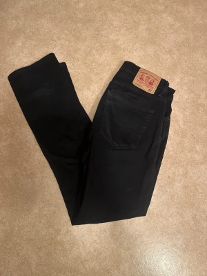 Svarta jeans - Säljer ett par svarta jeans 33/34 med klassisk femficksdesign och rak passform. Jeansen har en snygg läderpatch bak i midjan och är tillverkade i bomull med lite stretch för extra komfort. Perfekta för en clean och stilren look.