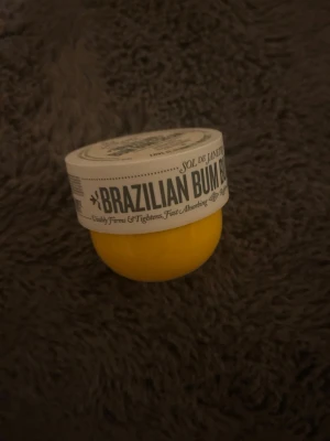 Sol de Janeiro Brazilian Bum Bum Cream - Ikonisk Brazilian Bum Bum Cream från Sol de Janeiro i en rund, gul burk med vitt lock. Krämen är känd för sin snabbt absorberande formula och doft av pistage och salt karamell. Perfekt för dig som vill ha en återfuktande och uppstramande kroppskräm.