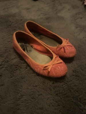 Korallfärgade ballerinaskor med rosett - Söta ballerinaskor i korallfärg med stickad struktur och liten rosett framtill. Skorna har rund tå och låg sula, perfekta för en avslappnad och trendig look. Insidan har ett blommigt mönster. Materialet ser ut att vara textil.