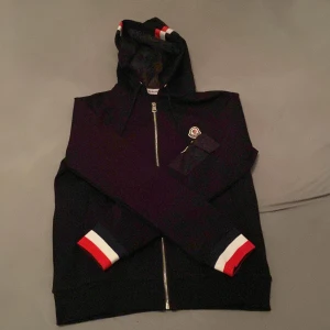Svart Moncler hoodie med dragkedja och byxor till - (Svarta moncler byxor medföljer)Svart hoodie från Moncler med hel dragkedja, huva och snygga rödvita detaljer på ärmslut och huva. Aldrig använd. Storlek M men sitter som S. Tröjan har Moncler-logga på bröstet och ärmen, samt ribbade muddar. Perfekt för dig som gillar stilrena streetwear-plagg med exklusiv känsla.