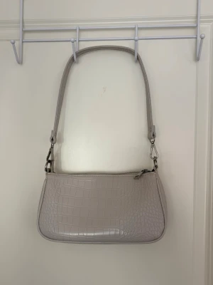 Beige axelväska - Stilren beige axelväska. Väskan har ett krokodil-mönster och stängs med dragkedja. Perfekt accessoar för att lyfta din outfit.