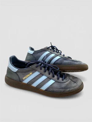 Adidas Spezial blå sneakers - Säljer ett par Adidas Spezial sneakers i grå mocka med ljusblå detaljer och klassiska tre ränder på sidan. Skorna har mörkblå snören, gummisula och guldfärgad Spezial-logga. Perfekt för dig som gillar retrostil och streetwear.