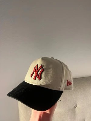Beige New York Yankees keps - Snygg beige keps från New Era med svart skärm och röd NY Yankees-logga framtill. Justerbar snapback baktill för perfekt passform. Broderad New Era-logga på sidan. Tillverkad i bomullstyg, perfekt för en sportig och trendig look.