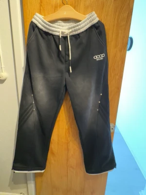 Svarta mjukisbyxor från Good Brand - Säljer ett par svarta mjukisbyxor från Good Brand med bred grå resår i midjan och vita dragsnören. Byxorna har raka ben, tryckknappar längs sidorna och två bakfickor. Perfekta för chill dagar eller träning.