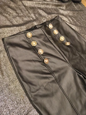 Svarta byxor med guldknappar - Snygga svarta byxor i skinnimitation med dekorativa guldknappar framtill. Hög midja och raka ben ger en trendig look. Perfekta för dig som vill sticka ut med coola detaljer och elegant finish.