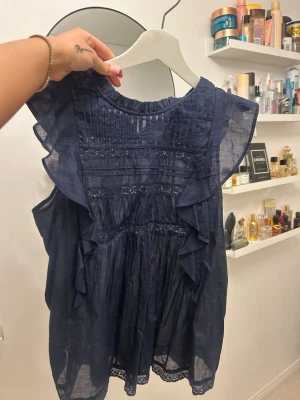 Volangblus  - Supersöt mörkblå blus med volanger längs axlarna och spetsdetaljer framtill och nedtill. Blusen är ärmlös och har en luftig, lätt transparent känsla. Perfekt till jeans eller kjol för en trendig look.