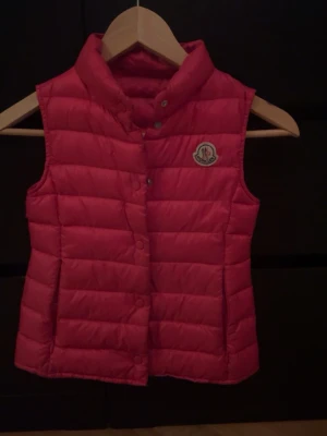 Rosa dunväst från Moncler - Snygg rosa dunväst från Moncler med quiltad design och hög krage. Västen har tryckknappar framtill och en broderad Moncler-logga på bröstet. Perfekt för lager-på-lager och håller dig varm med stil men även lägger till en snygg detalj u outfiten! Först i kvarn! 🌸🩷😁