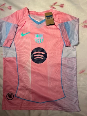 Barça rosa matchtröja Nike - Snygg rosa FC Barcelona matchtröja från Nike med ljusblå detaljer och klubbmärke på bröstet. Tröjan har V-ringning, korta ärmar och är tillverkad i ett lätt, ventilerande funktionsmaterial. Spotify-logga på magen och Nike swoosh i turkos.
