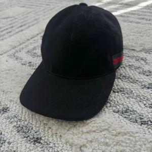 Svart keps från Gucci med rand äkta keps köpt från Gucci  - Snygg svart keps från Gucci med diskret GG-mönster i tyget och den klassiska gröna och röda randen på sidan. Kepsen har böjd skärm och justerbar rem bak. Perfekt för dig som vill ha en stilren accessoar med lyxig känsla äkta keps. 