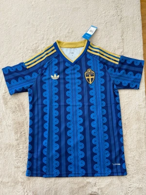Blå Sverige fotbollströja Adidas - Säljer en blå Sverige fotbollströja från Adidas med gul krage och gula ränder på axlarna. Tröjan har ett mönster i olika blå nyanser, broderad Adidas-logga och Sveriges landslagsemblem på bröstet. Materialet är lätt och andas, perfekt för träning eller match.