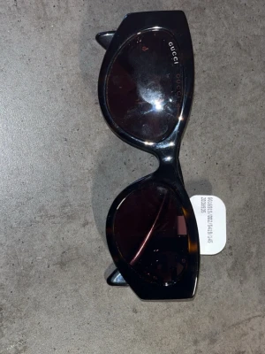 Svarta solglasögon från Gucci - Snygga svarta solglasögon från Gucci med bred båge och mörka glas. Klassisk cat-eye form och ikonisk guldig GG-detalj på skalmarna. Perfekta för dig som vill ha en lyxig och trendig accessoar till sommaren.