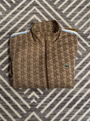 Brun mönstrad zip-tröja Lacoste - Snygg brun zip-tröja från Lacoste med geometriskt mönster och klassisk krokodil-logga på bröstet. Tröjan har höga krage och kontrastband i blått och vitt längs axlarna. Perfekt för dig som gillar sportig och trendig stil.
